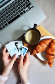 Hands are holding several instant photo prints next to a laptop, a cup of coffee, and two croissants on a plate. The setting appears to be an informal breakfast or coffee break scenario.