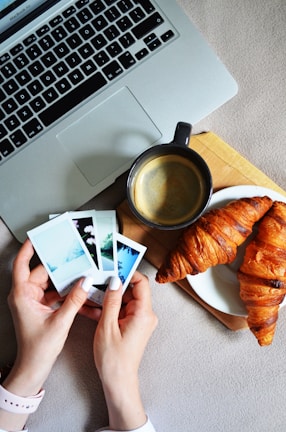 Hands are holding several instant photo prints next to a laptop, a cup of coffee, and two croissants on a plate. The setting appears to be an informal breakfast or coffee break scenario.
