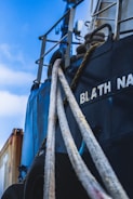 Detailed view of a ship with a close-up of its ropes and part of its hull. The ship's name is partially visible in bold white letters. The sky is blue with some clouds, and a cargo container is partially seen in the background.