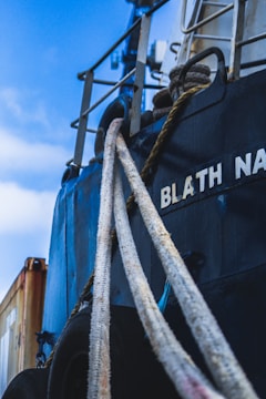 Detailed view of a ship with a close-up of its ropes and part of its hull. The ship's name is partially visible in bold white letters. The sky is blue with some clouds, and a cargo container is partially seen in the background.
