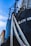 Detailed view of a ship with a close-up of its ropes and part of its hull. The ship's name is partially visible in bold white letters. The sky is blue with some clouds, and a cargo container is partially seen in the background.