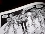 Three intricately drawn characters stand in conversation. Each has unique features and elaborate costumes with a variety of textures. The characters have expressive facial expressions and are engaged in a dialogue with speech bubbles indicating a lively exchange. The artwork is monochromatic, relying heavily on black and white shading.