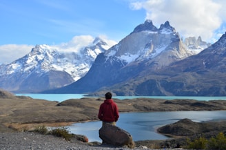 man in red hoodie sitting on rock near lake and snow covered mountain during daytime