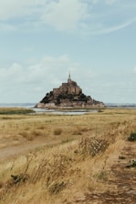 A historic island commune with a prominent abbey rising above the greenery and landscape, surrounded by vast stretches of grassy fields and a distant horizon under a partly cloudy sky.