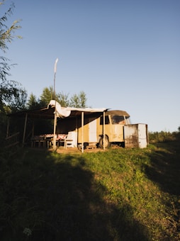 A rustic outdoor setting featuring an old, yellow camper van with a makeshift canopy attached to its side. The area around the van is grassy with some shadows cast by nearby trees. There is a wooden table with a checkered tablecloth under the canopy.