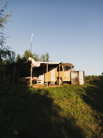 A rustic outdoor setting featuring an old, yellow camper van with a makeshift canopy attached to its side. The area around the van is grassy with some shadows cast by nearby trees. There is a wooden table with a checkered tablecloth under the canopy.