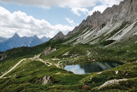 A picturesque landscape featuring a clear, still lake surrounded by lush green hills and rugged mountains. The sky is partly cloudy, with tall, jagged mountain peaks visible in the background. Small structures or huts are scattered across the landscape, connected by winding paths.