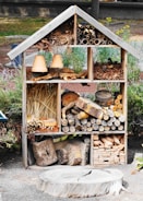 A wooden insect hotel is filled with various natural materials such as logs, pine cones, straw, and clay flower pots. The structure is divided into sections, each containing different items designed to attract and shelter insects. It is situated in an outdoor garden setting, surrounded by greenery and some visible fencing.