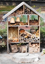A wooden insect hotel is filled with various natural materials such as logs, pine cones, straw, and clay flower pots. The structure is divided into sections, each containing different items designed to attract and shelter insects. It is situated in an outdoor garden setting, surrounded by greenery and some visible fencing.