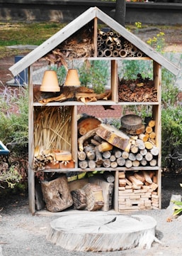 A wooden insect hotel is filled with various natural materials such as logs, pine cones, straw, and clay flower pots. The structure is divided into sections, each containing different items designed to attract and shelter insects. It is situated in an outdoor garden setting, surrounded by greenery and some visible fencing.