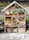 A wooden insect hotel is filled with various natural materials such as logs, pine cones, straw, and clay flower pots. The structure is divided into sections, each containing different items designed to attract and shelter insects. It is situated in an outdoor garden setting, surrounded by greenery and some visible fencing.