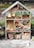 A wooden insect hotel is filled with various natural materials such as logs, pine cones, straw, and clay flower pots. The structure is divided into sections, each containing different items designed to attract and shelter insects. It is situated in an outdoor garden setting, surrounded by greenery and some visible fencing.