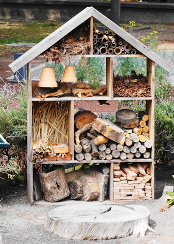 A wooden insect hotel is filled with various natural materials such as logs, pine cones, straw, and clay flower pots. The structure is divided into sections, each containing different items designed to attract and shelter insects. It is situated in an outdoor garden setting, surrounded by greenery and some visible fencing.