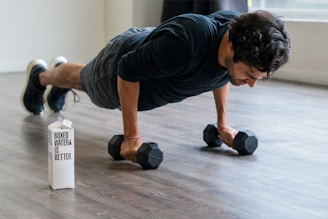 A person is performing a push-up exercise while gripping dumbbells on a wooden floor. Beside them, there is a box labeled 'Boxed Water Is Better'. The person is wearing a black long-sleeve shirt, gray shorts, and sneakers.