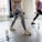 Two individuals are performing lunges in a well-lit studio, each holding dumbbells in their hands. They are both wearing athletic clothing, and cartons of boxed water are placed on the floor nearby. Large windows allow natural light to illuminate the wooden floor.