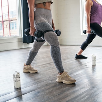 Two individuals are performing lunges in a well-lit studio, each holding dumbbells in their hands. They are both wearing athletic clothing, and cartons of boxed water are placed on the floor nearby. Large windows allow natural light to illuminate the wooden floor.