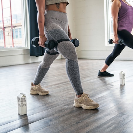 Two individuals are performing lunges in a well-lit studio, each holding dumbbells in their hands. They are both wearing athletic clothing, and cartons of boxed water are placed on the floor nearby. Large windows allow natural light to illuminate the wooden floor.