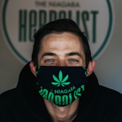 A person is wearing a black face mask with a green cannabis leaf design and the words 'The Niagara Herbalist'. Their eyes are visible above the mask and they have short hair, with a background featuring a logo or sign that is partially visible.