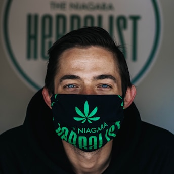 A person is wearing a black face mask with a green cannabis leaf design and the words 'The Niagara Herbalist'. Their eyes are visible above the mask and they have short hair, with a background featuring a logo or sign that is partially visible.