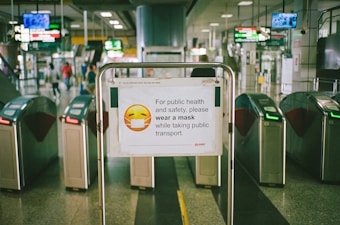 A sign with a masked emoji is displayed in a subway station, reminding passengers to wear masks for public health and safety. The surroundings include turnstiles and digital information boards displaying train schedules.