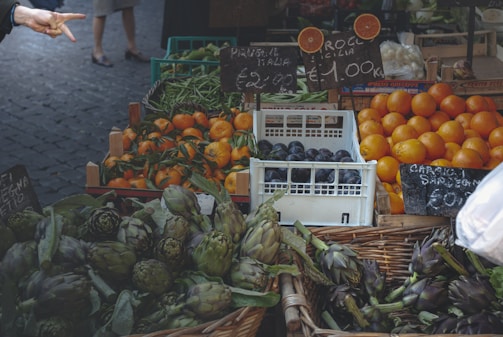 A vibrant outdoor market stand displaying a variety of fresh produce. The front baskets are filled with green artichokes and other leafy vegetables. To the right, there are stacks of oranges and purple plums in a white crate. Other visible produce includes green beans and oranges. Handwritten price signs are visible, indicating the cost per kilogram. A person's hand is pointing or making a gesture from the left side of the image.