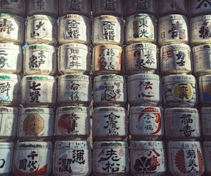 Rows of traditional Japanese sake barrels, intricately decorated with kanji characters and various colorful designs. The barrels are stacked closely together, creating a textured and cultural visual pattern.