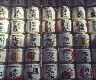 Rows of traditional Japanese sake barrels, intricately decorated with kanji characters and various colorful designs. The barrels are stacked closely together, creating a textured and cultural visual pattern.