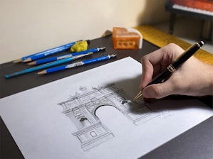 A hand drawing an architectural sketch of a triumphal arch on a sheet of paper. Surrounding the sheet are several blue pencils, a brush, and a pencil sharpener. The hand is holding a black and gold pen.