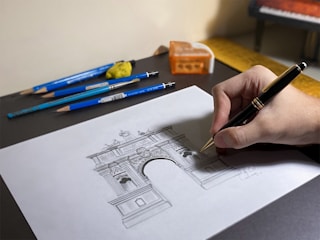 A hand drawing an architectural sketch of a triumphal arch on a sheet of paper. Surrounding the sheet are several blue pencils, a brush, and a pencil sharpener. The hand is holding a black and gold pen.