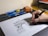 A hand drawing an architectural sketch of a triumphal arch on a sheet of paper. Surrounding the sheet are several blue pencils, a brush, and a pencil sharpener. The hand is holding a black and gold pen.