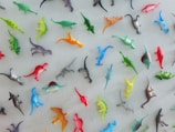 A collection of small, colorful plastic dinosaur figurines scattered on a flat surface. The dinosaurs are in various colors including green, blue, red, yellow, orange, and gray. They are arranged haphazardly with some facing different directions.