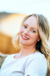 A woman with blonde hair and blue eyes is smiling, dressed in a white top with eyelet detailing. The soft, blurred background features warm tones, suggesting a sunny setting.