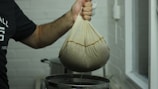A person is holding a cloth bag filled with a dark substance over a metal container. The bag appears to be used for straining or squeezing out liquid. The setting seems to be a kitchen or food preparation area with a light-colored brick wall in the background.