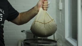 A person is holding a cloth bag filled with a dark substance over a metal container. The bag appears to be used for straining or squeezing out liquid. The setting seems to be a kitchen or food preparation area with a light-colored brick wall in the background.