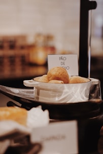 A small bowl filled with pão de queijo, a Brazilian cheese bread, is placed on a black serving stand. The background is softly blurred, highlighting the bowl and its contents. A sign next to the bowl clearly labels the item.
