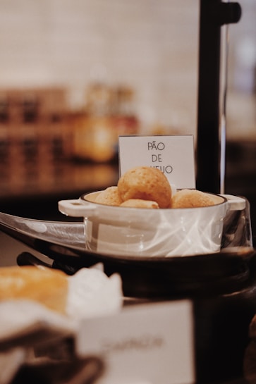 A small bowl filled with p&atilde;o de queijo, a Brazilian cheese bread, is placed on a black serving stand. The background is softly blurred, highlighting the bowl and its contents. A sign next to the bowl clearly labels the item.