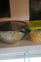 A large mixing bowl with a lid and a plastic container with a green lid are placed on a countertop. The container is filled with a mixture of corn, peas, and diced vegetables.