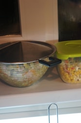 A large mixing bowl with a lid and a plastic container with a green lid are placed on a countertop. The container is filled with a mixture of corn, peas, and diced vegetables.