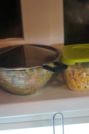 A large mixing bowl with a lid and a plastic container with a green lid are placed on a countertop. The container is filled with a mixture of corn, peas, and diced vegetables.