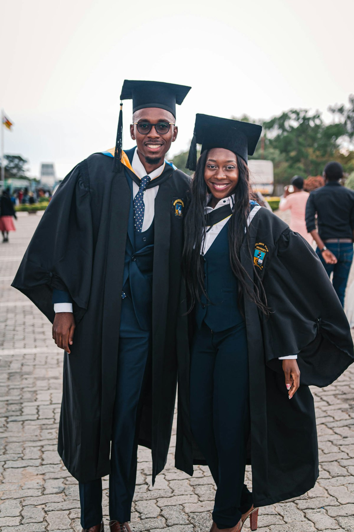 Graduate in black academic cap and gown standing in front of a university campus