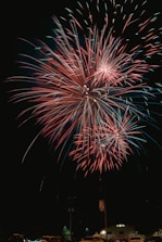 Bright and colorful fireworks burst in the night sky over a small area with vehicles and a building visible below. The fireworks display features vivid red and green hues creating a vibrant and celebratory atmosphere.
