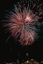 Bright and colorful fireworks burst in the night sky over a small area with vehicles and a building visible below. The fireworks display features vivid red and green hues creating a vibrant and celebratory atmosphere.