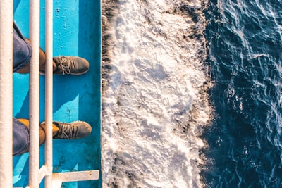 Footwear is visible from a top-down perspective on a blue metal surface adjacent to foamy water, indicating a scene of standing on a boat or ship.