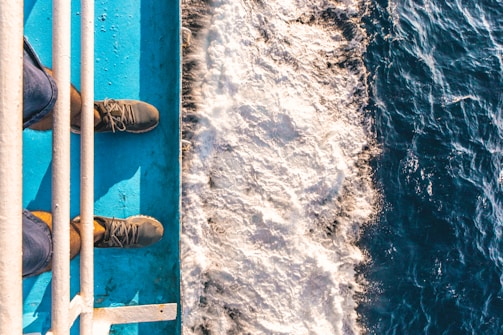 Footwear is visible from a top-down perspective on a blue metal surface adjacent to foamy water, indicating a scene of standing on a boat or ship.