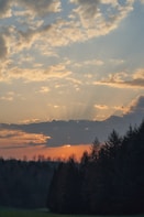 The image depicts a serene sunset over a forested landscape. The sky is filled with scattered clouds, allowing rays of sunlight to break through, creating a dramatic effect. Below, dark silhouettes of tall evergreen trees contrast against the warm colors of the sunset.