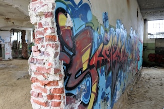 A dilapidated building interior with exposed brick and graffiti covering the walls. The room is filled with sand and debris, with colorful spray-painted art featuring bold strokes and overlapping colors.