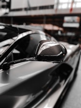 A close-up view of a sleek, glossy car side mirror with reflective surface. The background is blurred, suggesting an indoor setting with ambient lighting and what appears to be showroom elements.