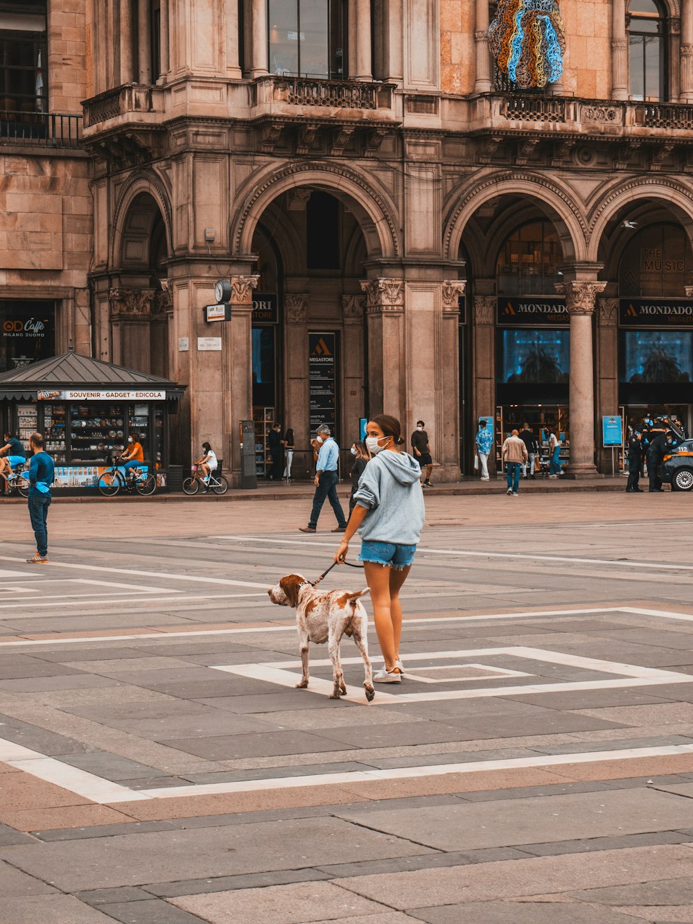 woman in teal shirt and blue denim shorts walking with brown dog on pedestrian lane during