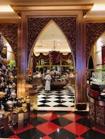 An ornate and luxurious interior space featuring a chef standing near a circular counter. The floor is decorated with black and white checkerboard tiles transitioning to red and black tiles. Elegant gold and maroon arches frame the scene. A large Christmas tree adorned with golden ornaments and presents is visible on the left.
