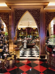 An ornate and luxurious interior space featuring a chef standing near a circular counter. The floor is decorated with black and white checkerboard tiles transitioning to red and black tiles. Elegant gold and maroon arches frame the scene. A large Christmas tree adorned with golden ornaments and presents is visible on the left.